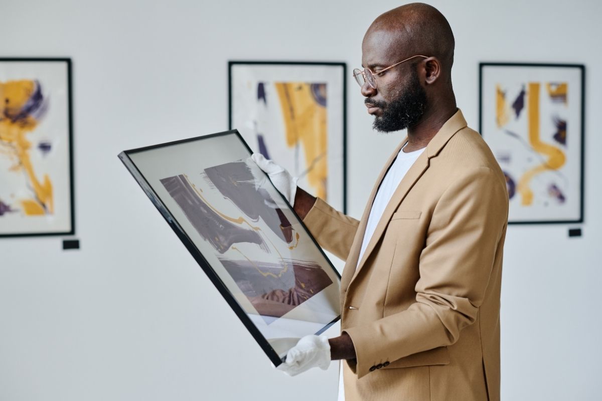 A gallerist wearing gloves examining a framed abstract artwork in a gallery