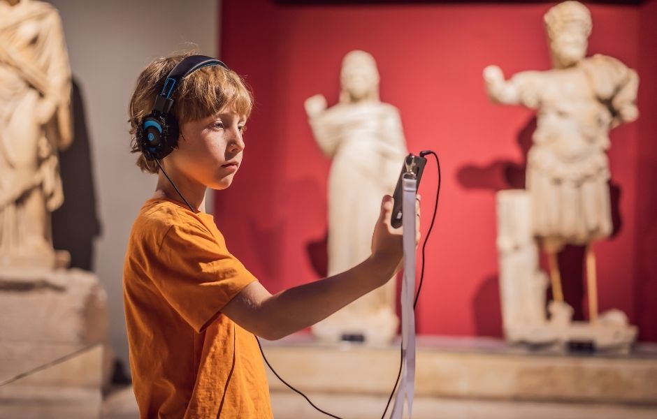 Child using an audio guide and handheld device among classical sculptures, representing the diverse media collections institutions manage with Gallery Systems