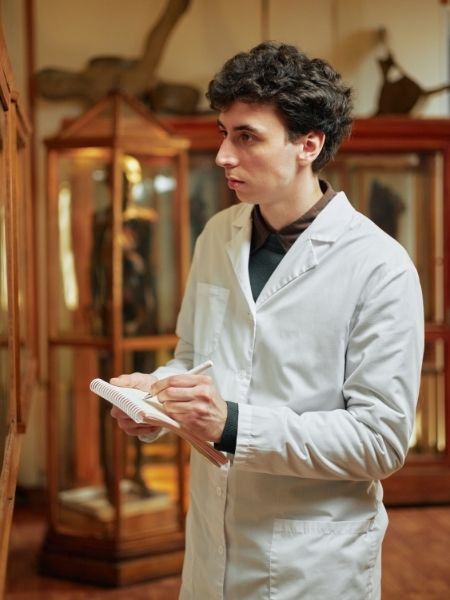 A conservator in a white lab coat taking notes while examining objects in a natural history gallery, representing the collections management workflows TMS Conservation Studio is designed to support