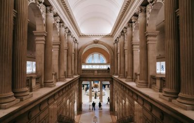The grand interior hall of a major cultural institution with towering classical columns, vaulted ceilings, and visitors visible on the lower level