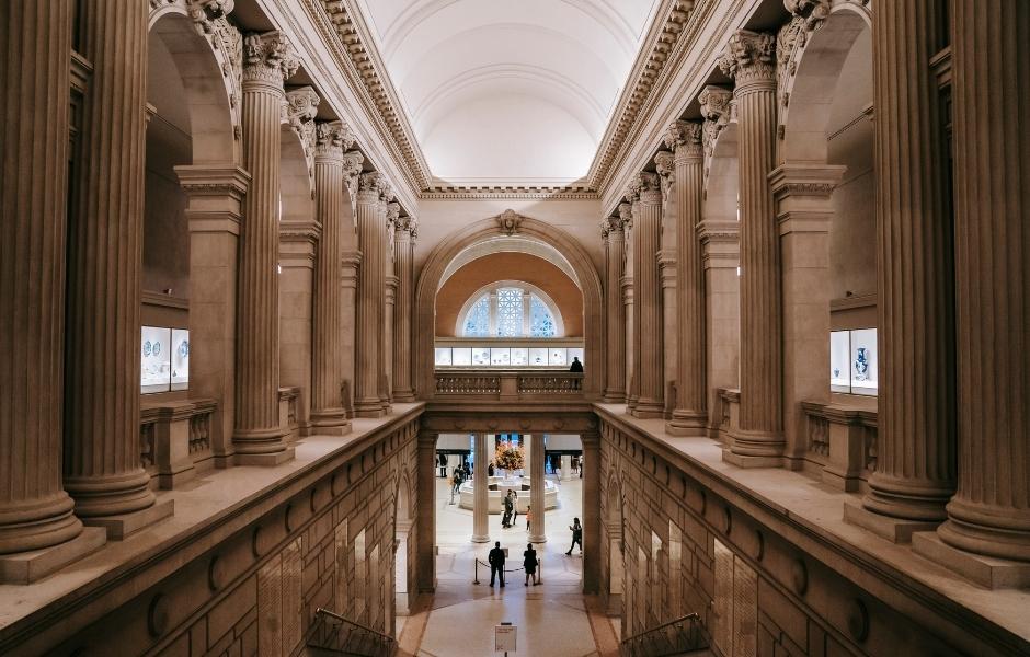The grand interior hall of a major cultural institution with towering classical columns, vaulted ceilings, and visitors visible on the lower level