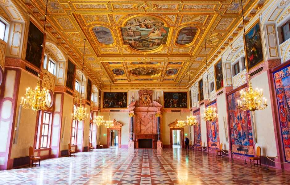 A grand institutional hall with a painted ceiling, gold chandeliers, and large tapestries lining the walls