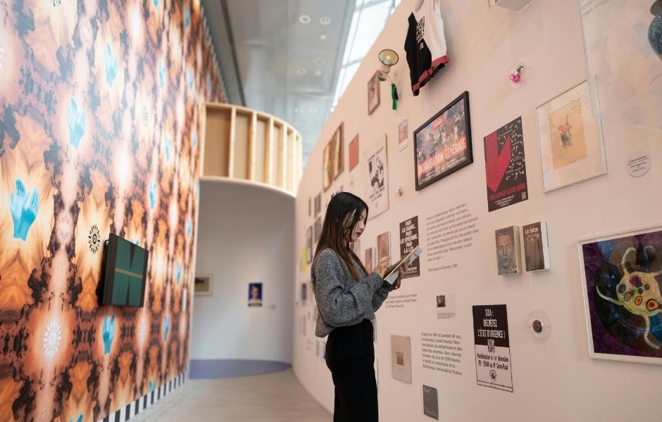 A visitor consulting a mobile device in front of a densely curated exhibition wall displaying mixed media works and printed materials
