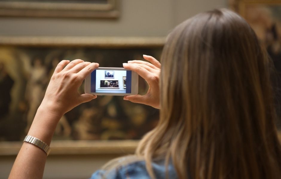 A visitor photographing framed paintings in a gallery on a smartphone, capturing digital assets linked to collection records