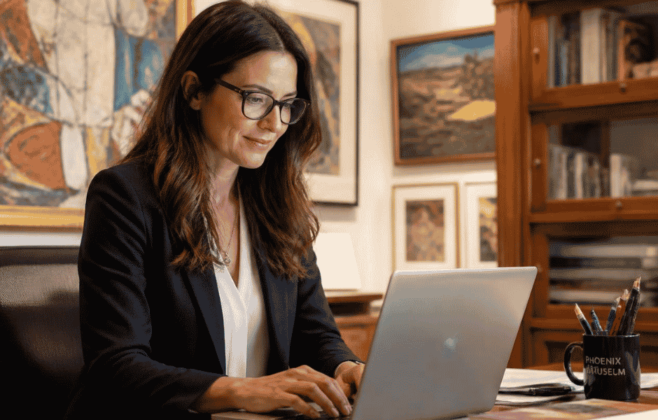 A collections professional working at a laptop in an office surrounded by framed artworks, representing individual staff access to a unified collections management system