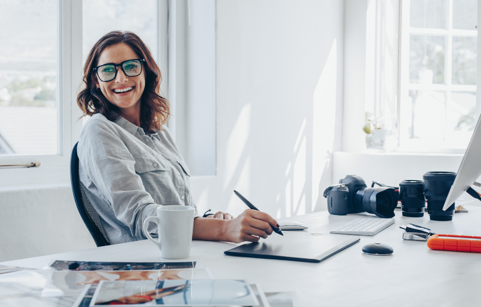 A smiling woman with shoulder-length brown hair and glasses sits at a bright, modern desk. She is using a digital pen on a graphic tablet, with a white coffee mug, photographs, a camera, camera lenses, and a computer monitor visible on the desk. Sunlight streams through large windows in the background, creating a cheerful and creative workspace atmosphere.