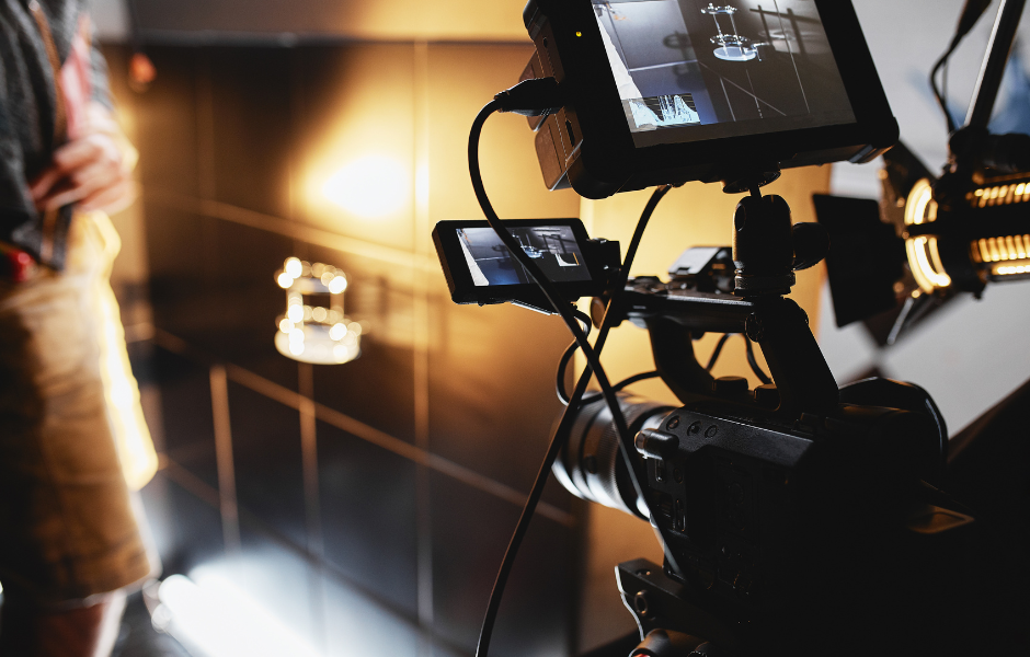 A close-up view of a professional video camera setup on a film set, with external monitors displaying a live feed of the scene being filmed. The scene includes a suspended object under focused lighting. The background shows reflective black tiles and warm studio lighting, with part of a crew member visible on the left side of the frame.