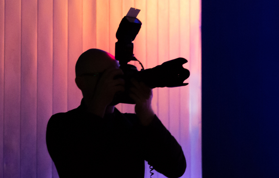 A silhouette of a photographer holding a professional DSLR camera with an external flash attachment, set against a vibrant background of vertical blinds illuminated by pink, purple, and blue lighting. The scene conveys a dramatic and creative atmosphere, with the photographer's features obscured in shadow.